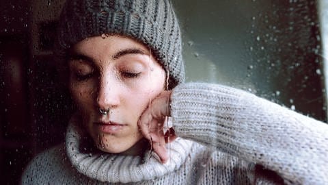 Junge Frau mit geschlossenen Augen steht hinter einer verregneten Fensterscheibe. (Foto: IMAGO, IMAGO / Addictive Stock)