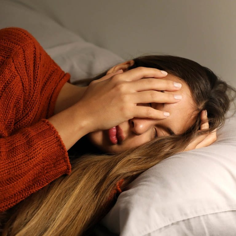 Junge Frau im Bett hält sich das Gesicht mit den Händen zu. (Foto: IMAGO, IMAGO / Panthermedia)