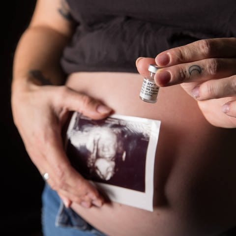 Frau hält Ultraschallbild und Impfampulle vor ihren schwangeren Bauch. (Foto: IMAGO, IMAGO / Fotostand)
