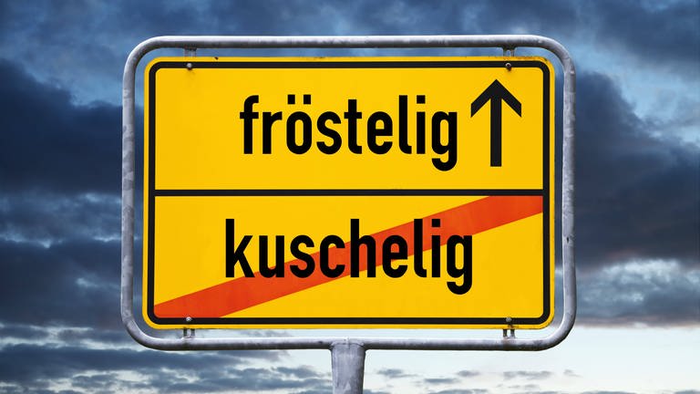 Ortsschild mit Aufschrift "fröstelig" und durchgestrichener Aufschrift "kuschelig". (Foto: IMAGO, IMAGO / Christian Ohde)