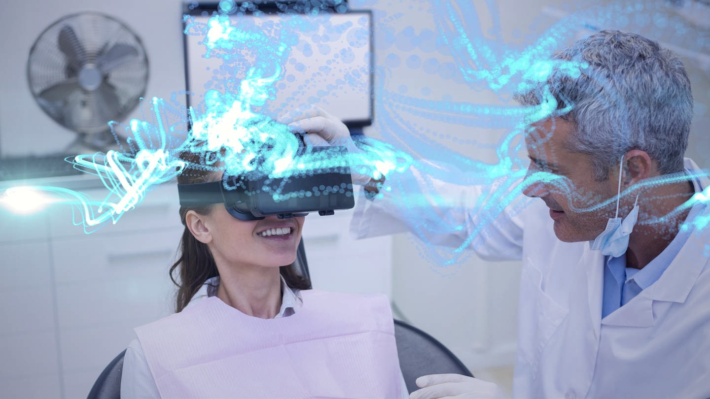 Eine Frau auf einer Behandlungsliege trägt eine VR-Brille und lacht, während ein Mann den Sitz der VR-Brille bei ihr überprüft. (Foto: IMAGO, -)