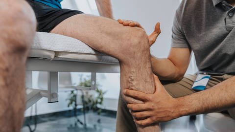 Physiotherapeut berührt das Knie eines männlichen Patienten. (Foto: IMAGO, IMAGO / Westend61)