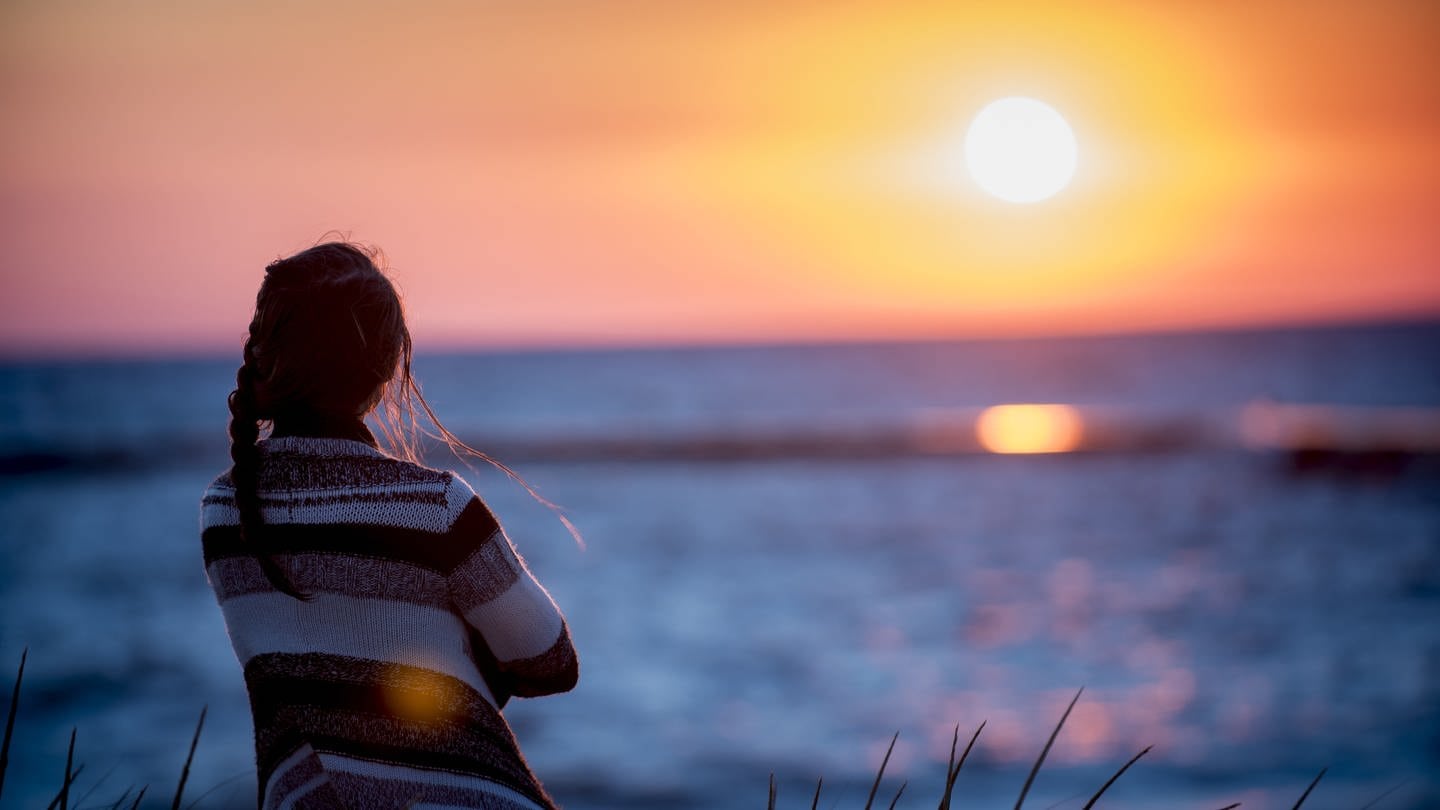 Eine Frau beobachtet den Sonnenaufgang über dem Meer. (Foto: IMAGO, IMAGO / Wirestock)