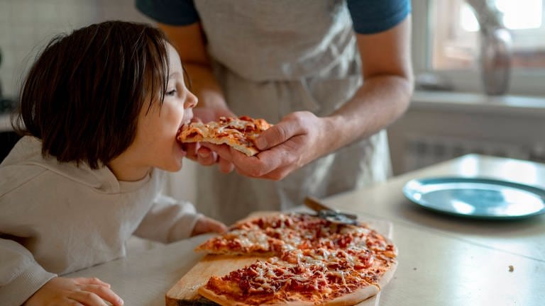 Kind wird von dem Vater mit einer Pizza gefüttert. (Foto: IMAGO, /Westend61)