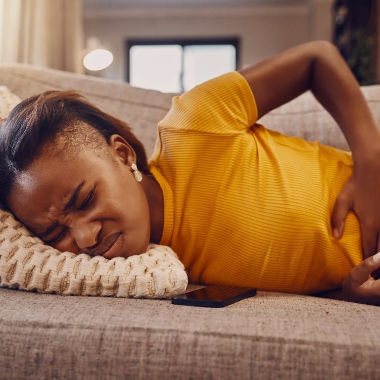 Eine Frau mit Bauchschmerzen und schmerzverzerrtem Gesicht liegt auf einem Sofa und hält sich den Bauch. (Foto: IMAGO, IMAGO / Zoonar)
