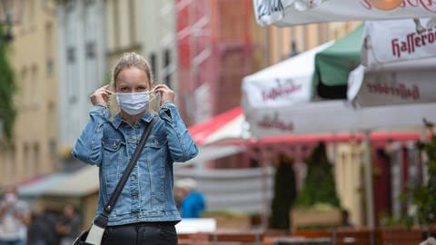 Frau vor Biergarten in einer Stadt zieht sich eine Maske an (Foto: IMAGO, imago images / MedienServiceMüller)