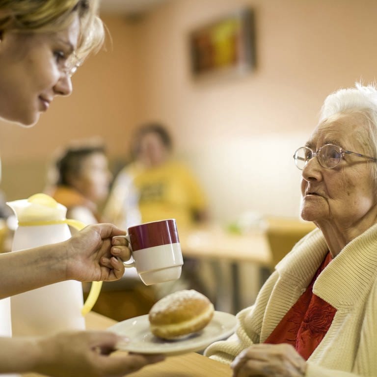 Pflegerin reicht einer alten Dame eine Tasse (Foto: IMAGO, imago/allOver-MEV)