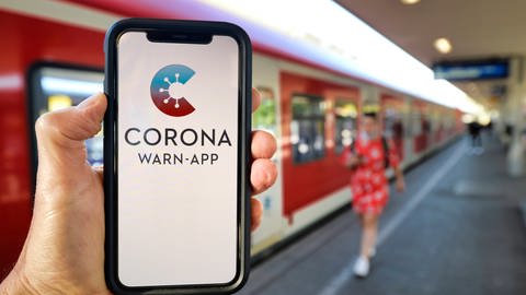 Hand hält Smartphone mit Corona-Warn-App am Bahnhof (Foto: IMAGO, imago images / Michael Weber)