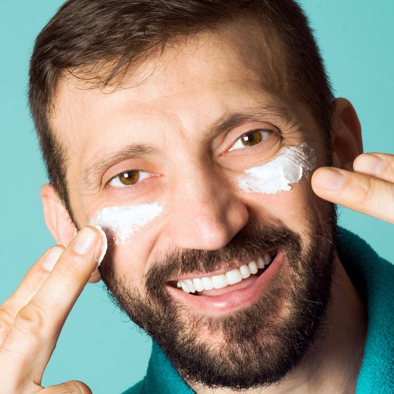 Ein Mann streicht Creme auf seine Augenringe und lacht (Foto: IMAGO, NikoletaVukovic via www.imago-images.de)