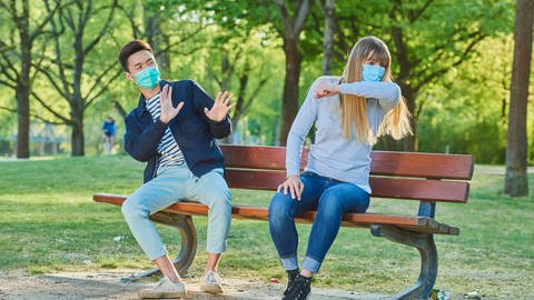 Mann und Frau mit Mundschutzmaske auf einer Parkbank in einem Stadtpark. (Symbolbild) (Foto: IMAGO, imago/ imageBROKER/David&Micha Sheldon)