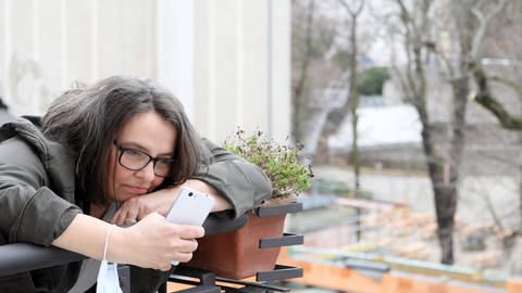 Frau auf einem Balkon am Handy mit Maske am Unterarm