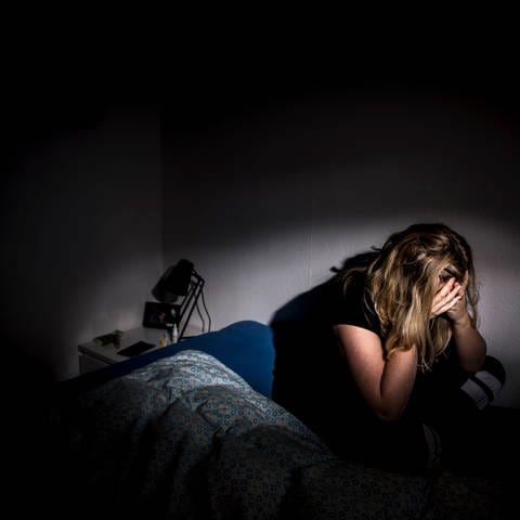 Eine Frau sitzt, das Gesicht in den Händen vergraben, in einem dunklen Raum auf einem Bett.