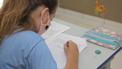 Mädchen mit Stoffmaske sitzt über ihren Hausaufgaben (Foto: IMAGO, Imago Images / Fotoarena)