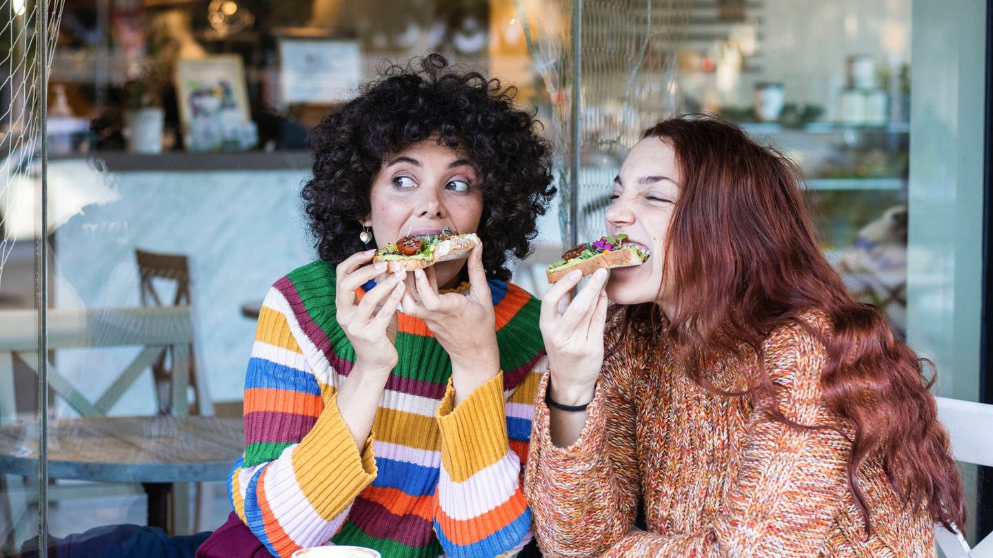 Zwei Frauen beißen beim Frühstück herzhaft in belegte Brote. (Foto: IMAGO, IMAGO / Westend61)