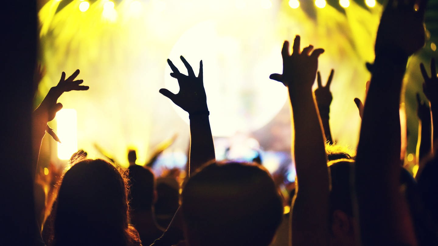 Menschen heben vor einer beleuchteten Bühne die Hände zum Feiern in die Höhe. (Foto: IMAGO, IMAGO / agefotostock)