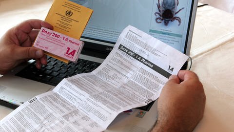 Betroffener sitzt mit Impfpass, Medikament Doxy 200 und Beipackzettel vor einer Internetseite zur Zecke (Foto: IMAGO, / suedraumfoto)