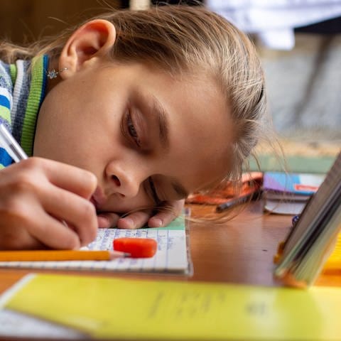 Kind lehnt kopf auf Schreibtisch und schreibt. (Foto: IMAGO, IMAGO / Shotshop)