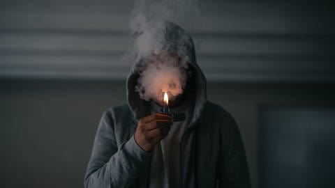 Mann zündet sich einen Joint an, das Gesicht verbirgt sich hinter einer Rauchwolke. (Foto: IMAGO, IMAGO / Addictive Stock)