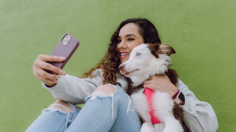 Zufriedener weiblicher Besitzer, der ein Selbstporträt mit seinem Border Collie-Hund auf seinem Smartphone aufnimmt, während er in der Nähe einer grünen Wand in einer Stadtstraße sitzt.
