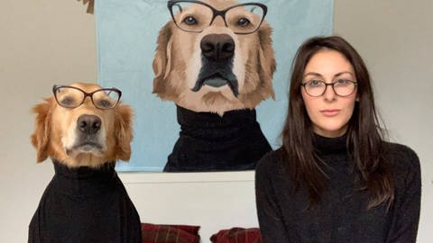 Frau und Hund sind gleich angezogen mit schwarzem Rollkragenpulli und Brille, wie ein Bild, was im Hintergrund zu sehen ist.
