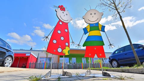 Strichmännchen-Figuren von einem Mädchen und einem Jungen stehen vor einem Kindergarten.