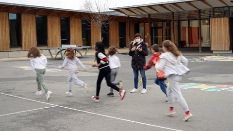 Schülerinnen rennen Runden vor dem Schulhof. Eine Lehrerin mit Maske misst die Zeit. (Foto: IMAGO, IMAGO / Hans Lucas)