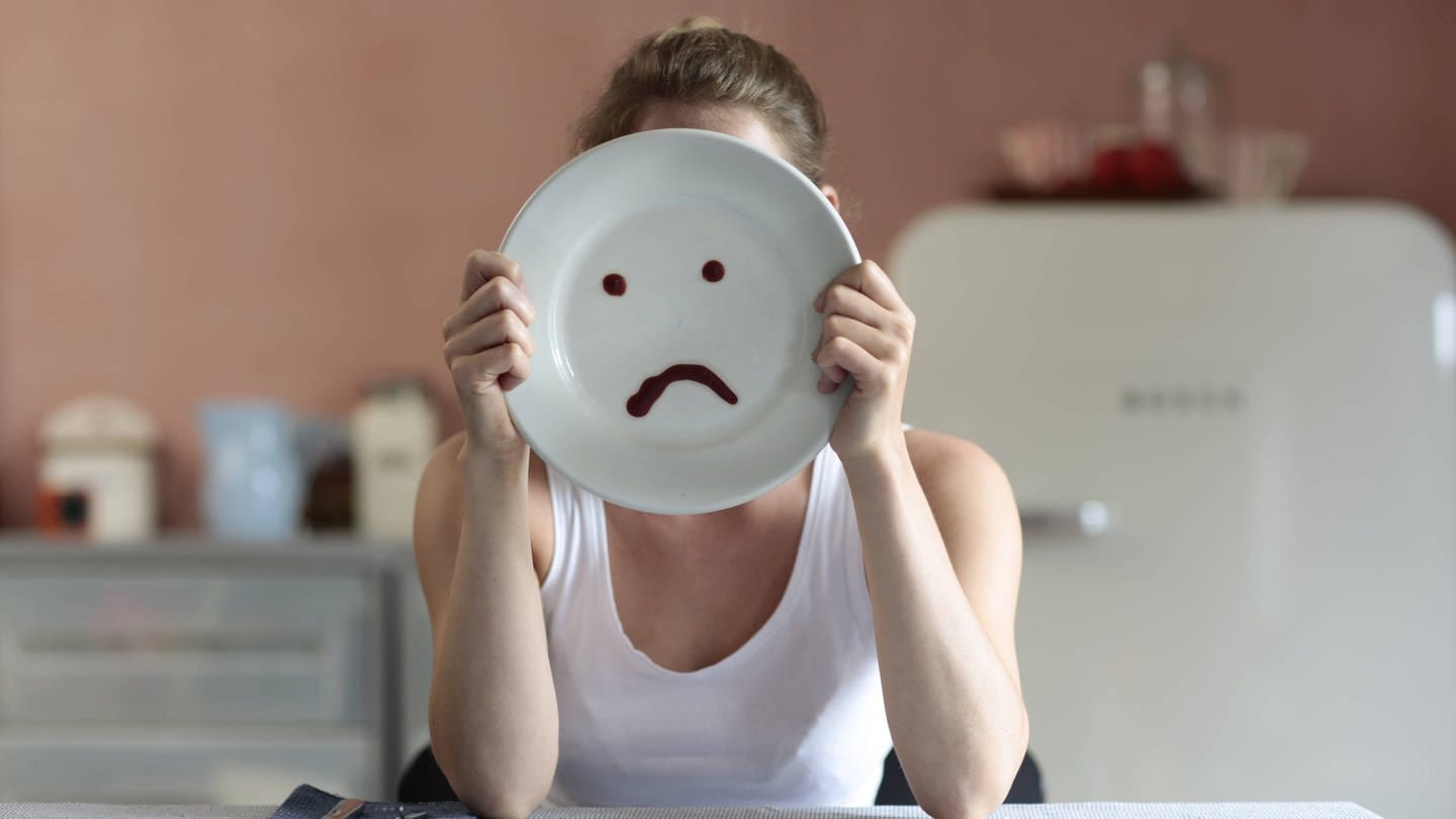 Neue Forschung zeigt: Dlk1-Neuronen im Gehirn steuern Appetitlosigkeit. (Symbolbild: Man sieht eine Frau, die sich einen Teller vors Gesicht hält, auf dem ein trauriger Smiley zu sehen ist.) (Foto: IMAGO, photothek)