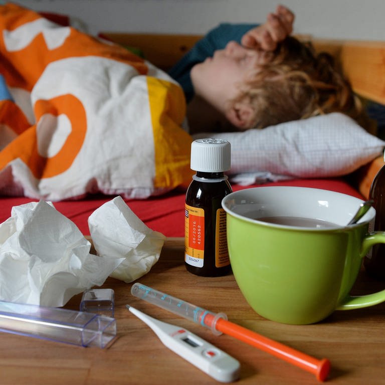 Ein Junge liegt mit Fieber im Bett. Vor ihm auf dem Tisch steht ein Fiebersaft