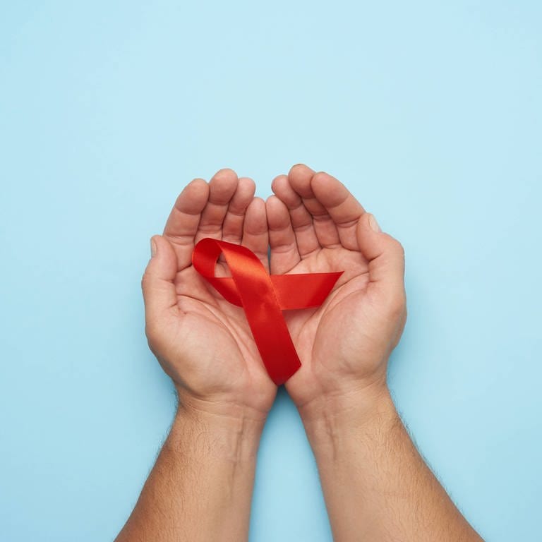 Hände eines Mannes halten eine rote Schleife - ein Symbol für den Kampf gegen AIDS. (Foto: IMAGO, IMAGO / YAY Images)