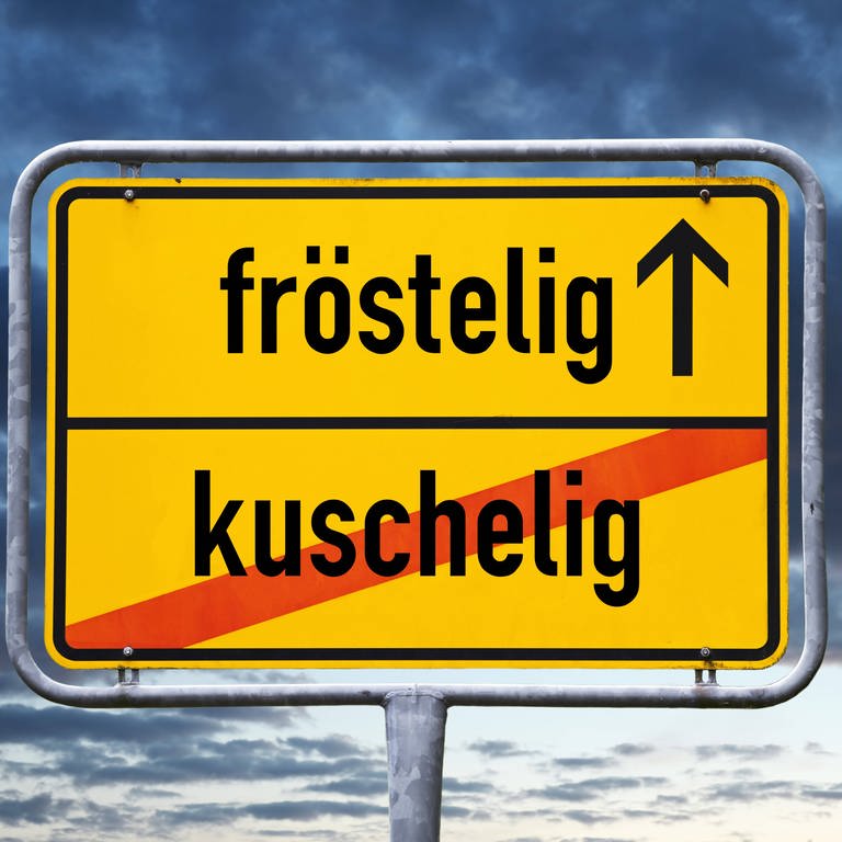 Ortsschild mit Aufschrift "fröstelig" und durchgestrichener Aufschrift "kuschelig". (Foto: IMAGO, IMAGO / Christian Ohde)