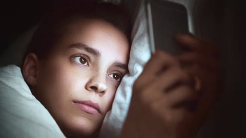 Frau liegt im Bett und schaut auf ihr Handy. (Foto: IMAGO, IMAGO / agefotostock)