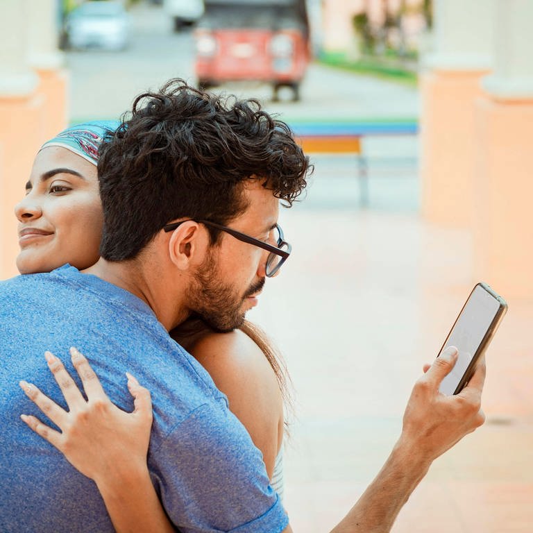 Mann schaut auf sein Handy während er seine Freundin umarmt. 