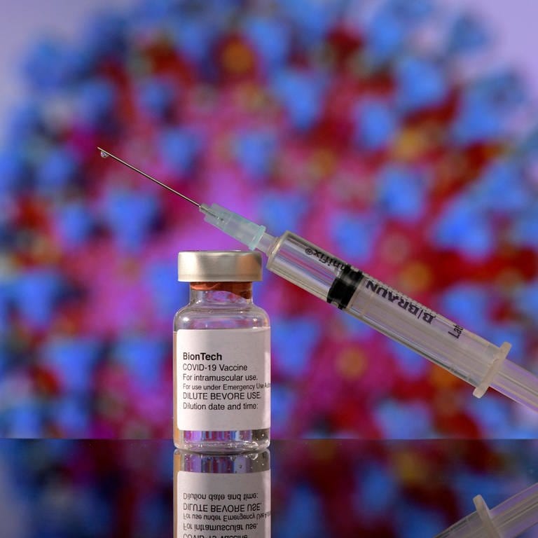 Eine Biontech-Ampulle und Spritze liegen auf dem Tisch. Im Hintergrund ist ein großes Coronavirus Symbol zu sehen. (Foto: IMAGO, IMAGO / MiS)