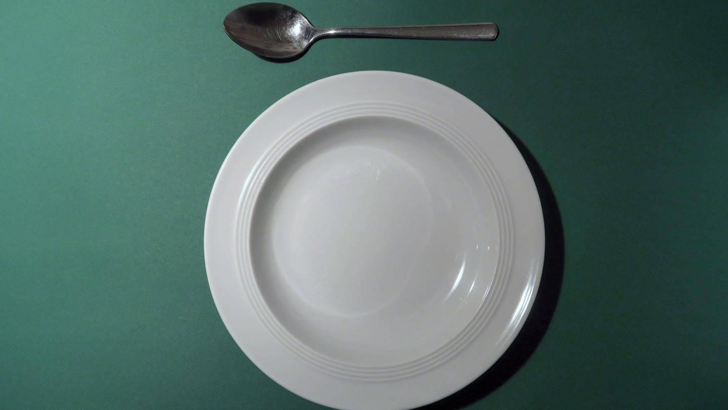 Ein leerer Teller und ein Löffel liegen auf einer grünen Unterlage. (Foto: IMAGO, IMAGO / imagebroker)