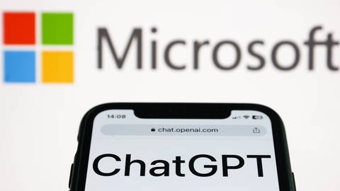 Das Bild zeigt ein Handy, auf dessen Bildschirm ChatGPT angezeigt wird und im Hintergrund befindet sich das Microsoft-Logo.