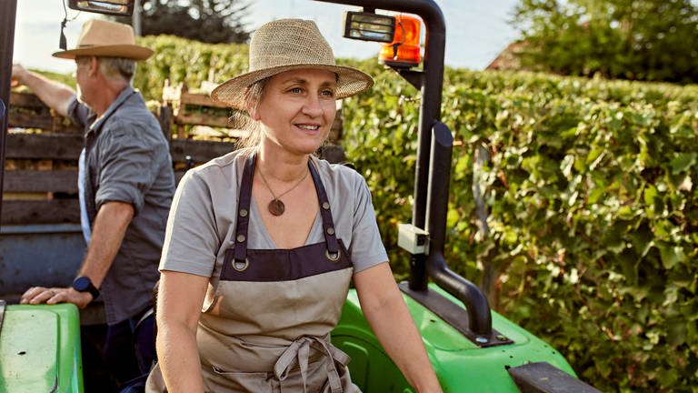 Eine lächelnde Frau fährt einen Traktor durch auf einem Acker, hinter ihr sitzt ein Kollege (Foto: IMAGO, Westend61)