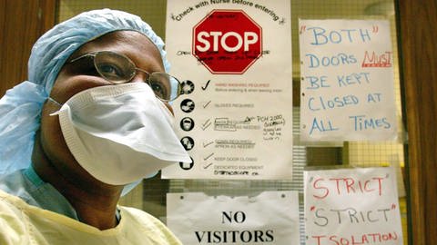 ine Krankenschwester trägt Schutzkleidung gegen SARS. Im Hintergrund kleben Warnhinweise zu Hygienemaßnahmen.