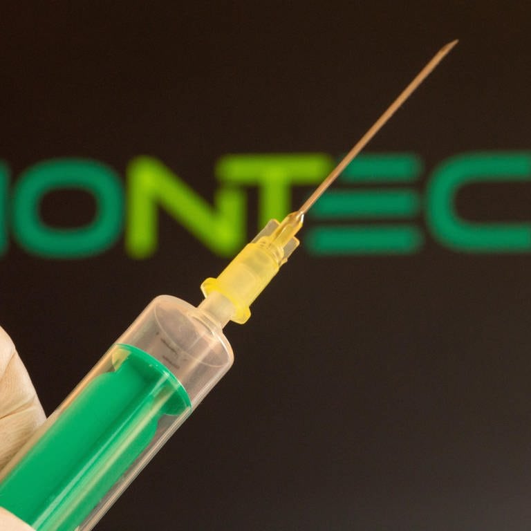 Nahaufnahme einer Injektionsnadel, im Hintergrund das Logo von Biontech. Die Debatte über mögliche Impfschäden nimmt in Deutschland derzeit an Fahrt auf