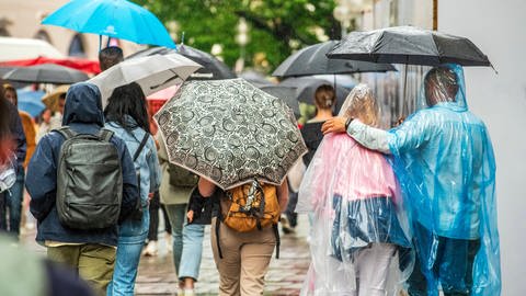 Menschen in Fußgängerzone schützen sich mit Regenschirmen und -capes vor dem Regen.