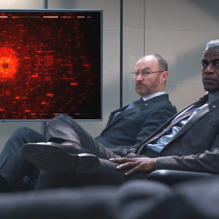 Aufnahme aus dem Film: Zwei Männer sitzen vor einem Bildschirm, auf dem die KI abstrakt abgebildet ist 