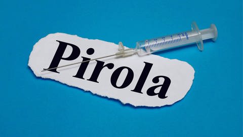 Das Bild zeigt eine Impfspritze vor einem Zettel mit der Aufschrift "Pirola".