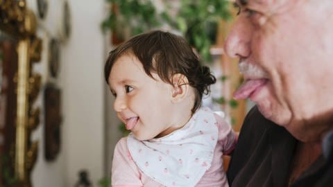 Das Bild zeigt ein kleines Mädchen auf dem Arm eines älteren Mannes, die beide ihre Zungen rausstrecken. Symbolbild.