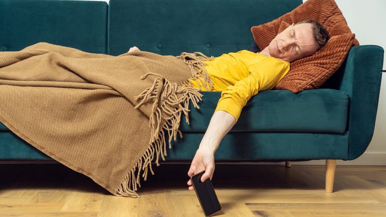 Müder Mann schläft auf Sofa. Sein Arm hängt auf den Boden und hält sein Smartphone in der Hand. Laut Studie schlafen wir kürzer.