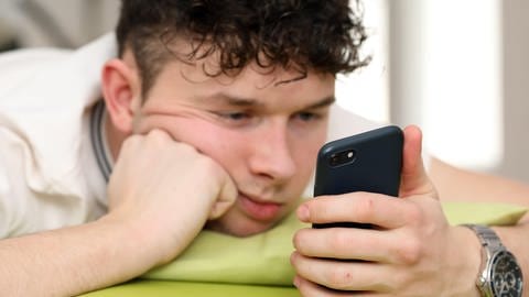 Teenager schaut im Bett gelangweilt auf sein Smartphone. Jugendliche haben meist noch keine ausreichende Selbstregulation und nutzen Social Media daher exzessiver.