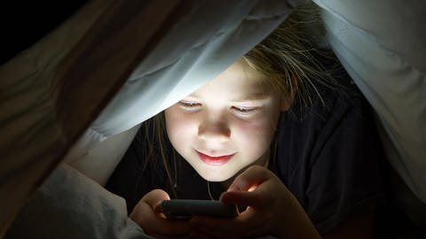 Mädchen sitzt unter Bettdecke und schaut auf ein Handy.