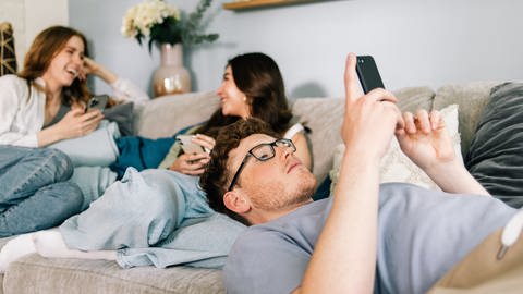 Mehrere Teenager sitzen im Wohnzimmer und schauen auf ihre Smartphones. Soziale Medien können ein Gemeinschaftsgefühl schaffen, aber auch die Flucht aus dem Alltag unterstützen.