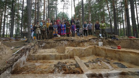 Das Grabungsteam und ortsansässige indigene Partner aus der Gruppe der Chanten stehen vor einem Grabungschnitt in Kayukovo. Im hellen Sand zeichnen sich dunkel die Reste eines verbrannten steinzeitlichen Hauses ab.  (Foto: Sventlana Lips)
