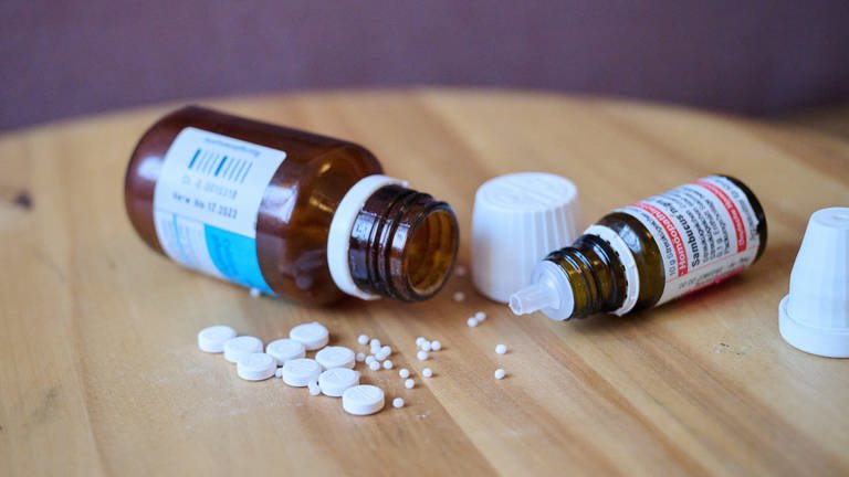 Homöopathische Glasfläschchen, Kügelchen und Tabletten liegen auf einem Tisch. Ein Kommentar dazu, dass Homöopathie nicht länger Kassenleistung bleiben soll.