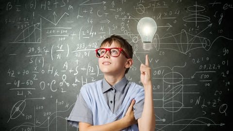 Junge steht vor Klassentafel und zeigt auf Glühbirne über seinem Kopf.