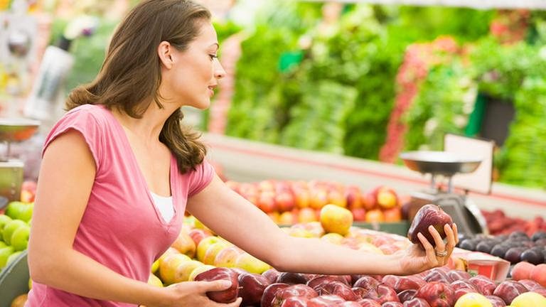 Eine Frau beim Einkaufen, sie hält einen roten Apfel in der Hand und begutachtet ihn.